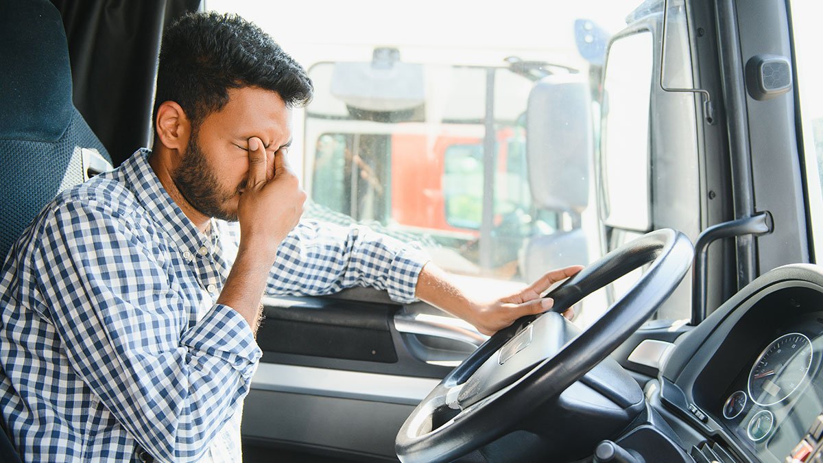 A tired truck driver in a plaid shirt pinching the bridge of his nose while resting at the wheel of his semi truck