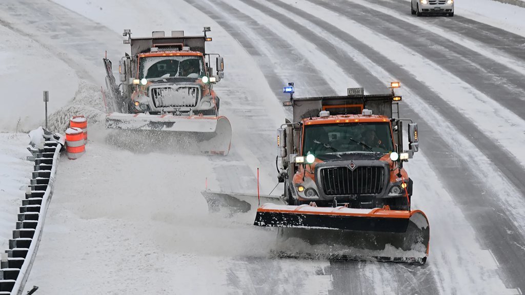 Two orange snow plow trucks with amber lights clearing snow from a highway during a Minnesota winter storm