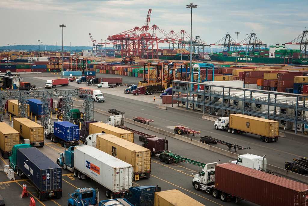 Aerial view of a container port terminal with red gantry cranes, stacked containers, and semi trucks hauling chassis